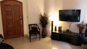Un salon avec une porte en bois, un fauteuil noir, un grand vase contenant des plantes séchées, un téléviseur posé sur un meuble et une lampe.