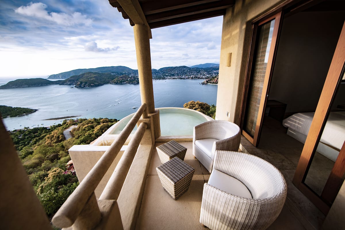A balcony with a hot tub, wicker furniture, and a view of a lake and hills.