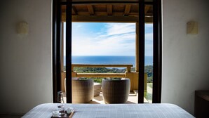 Un dormitorio con una cama, dos sillas de mimbre, una mesa con vasos y una bandeja, y un balcón con vistas al mar y al verdor.