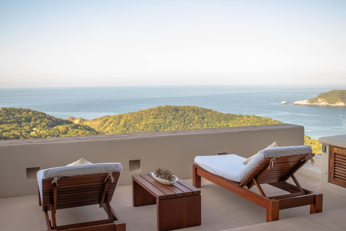 A balcony with two wooden lounge chairs and a small table, overlooking a coastal landscape with greenery and the sea.