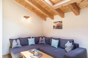 Une pièce avec un plafond en bois, un canapé gris agrémenté de plusieurs coussins, une petite table avec un verre et un luminaire fixé au mur.