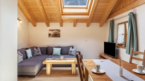 Ein gemütliches Zimmer mit Holzbalkendecke, einer Couch, einem Esstisch und einem Fernseher.