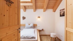 Ein Zimmer mit Holzboden, einem Bett, einem kleinen Hocker, einer Wandleuchte und einer Holzzieldecke.