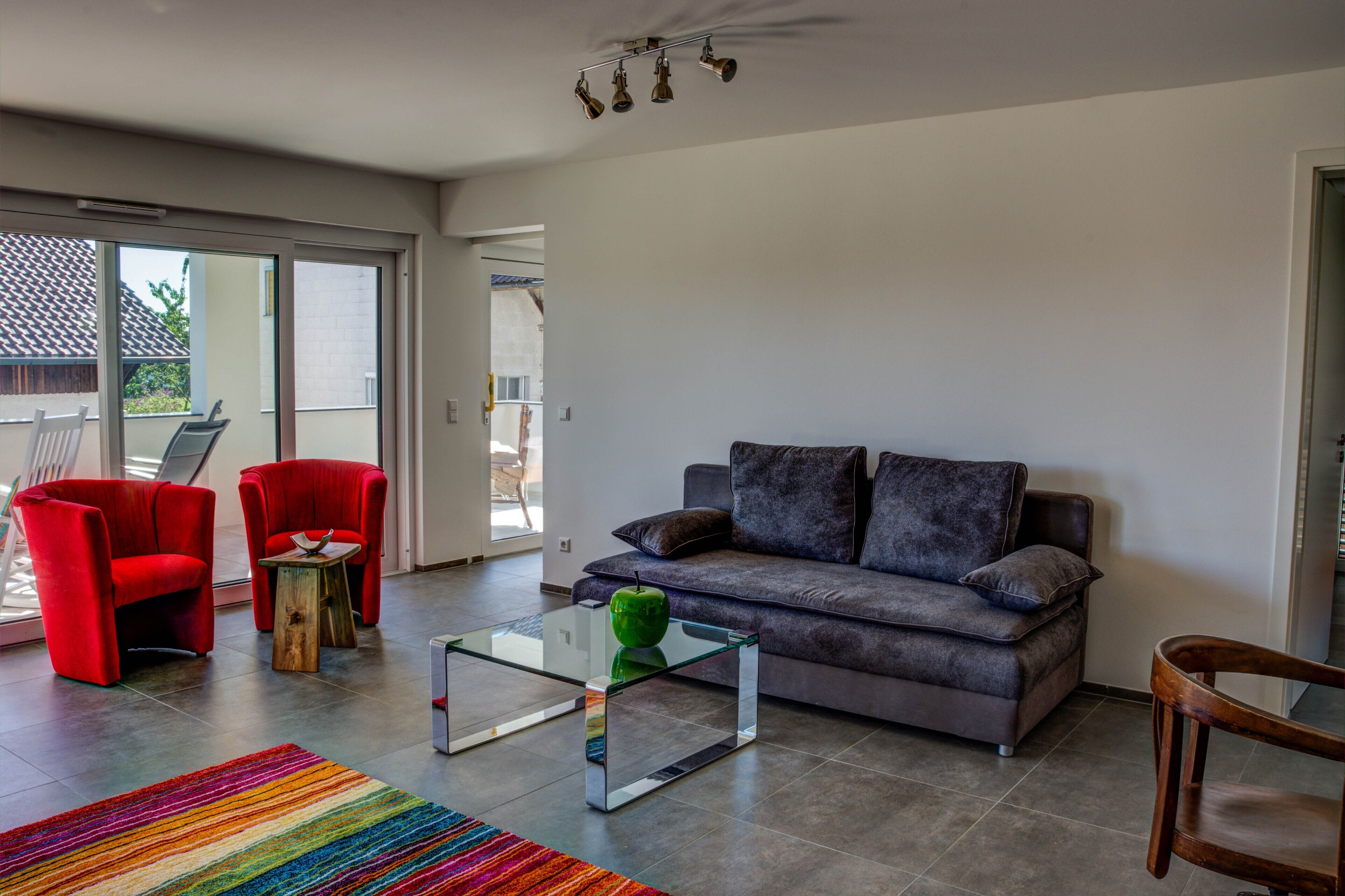 Ein Wohnzimmer mit grauem Sofa, roten Sesseln, einem Glastisch und einem bunten Teppich.