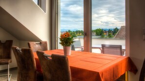 A dining area with a table and chairs, a potted plant, and a view through large windows.