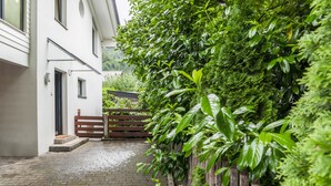 A paved courtyard with a wooden gate, a white building with a door and windows, and dense green foliage. - Vacation Home "Das Bergzeit" with Mountain View, Wi-Fi, Garden & Terrace (Farchant)