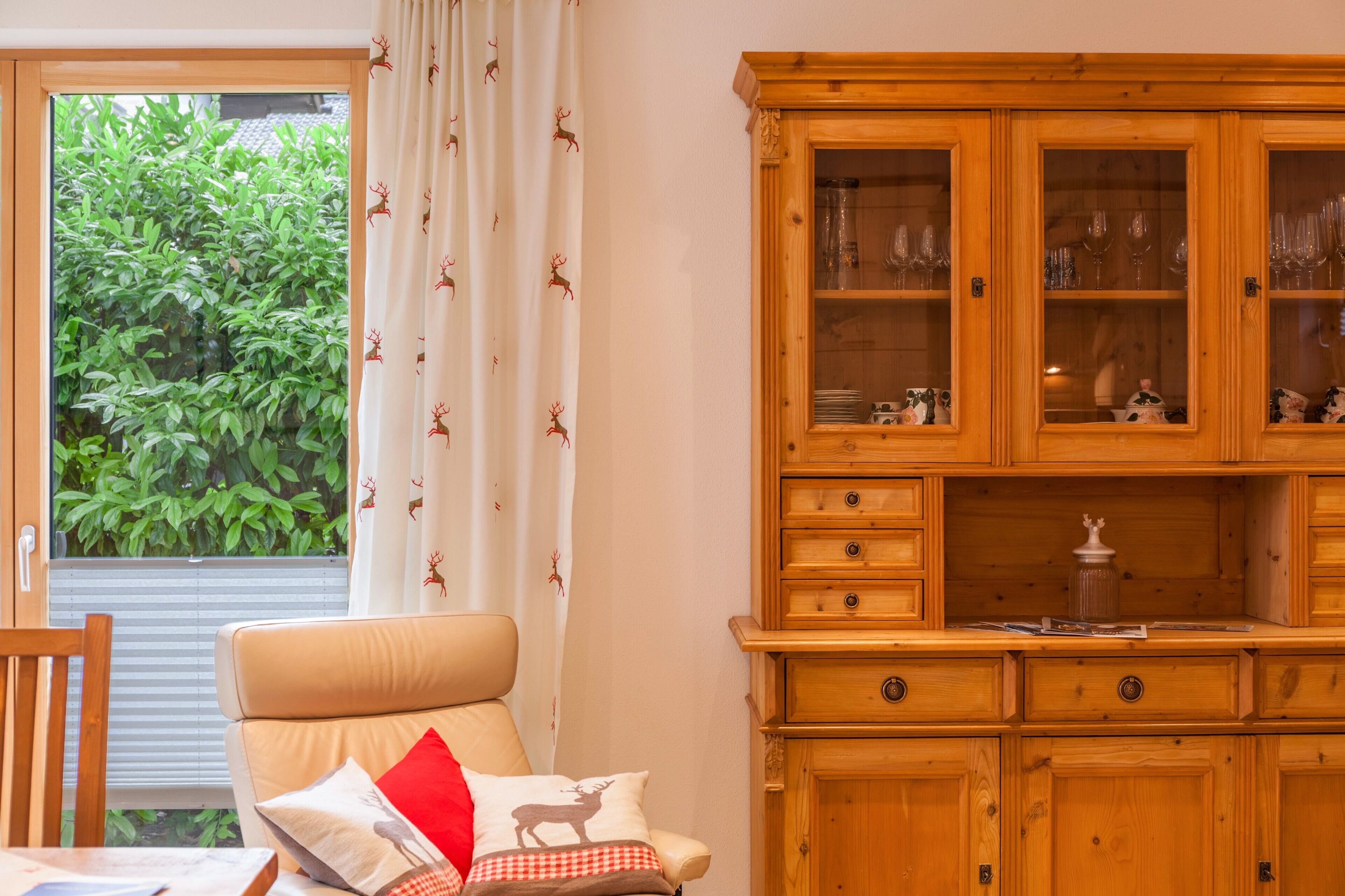 Ein Zimmer mit Fenster, einem Holzschrank und einem Sessel mit Kissen.