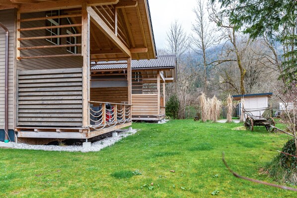 Une cabane en bois avec un porche, une charrette et un petit abri, située dans une zone herbeuse, avec des arbres et des montagnes en arrière-plan.