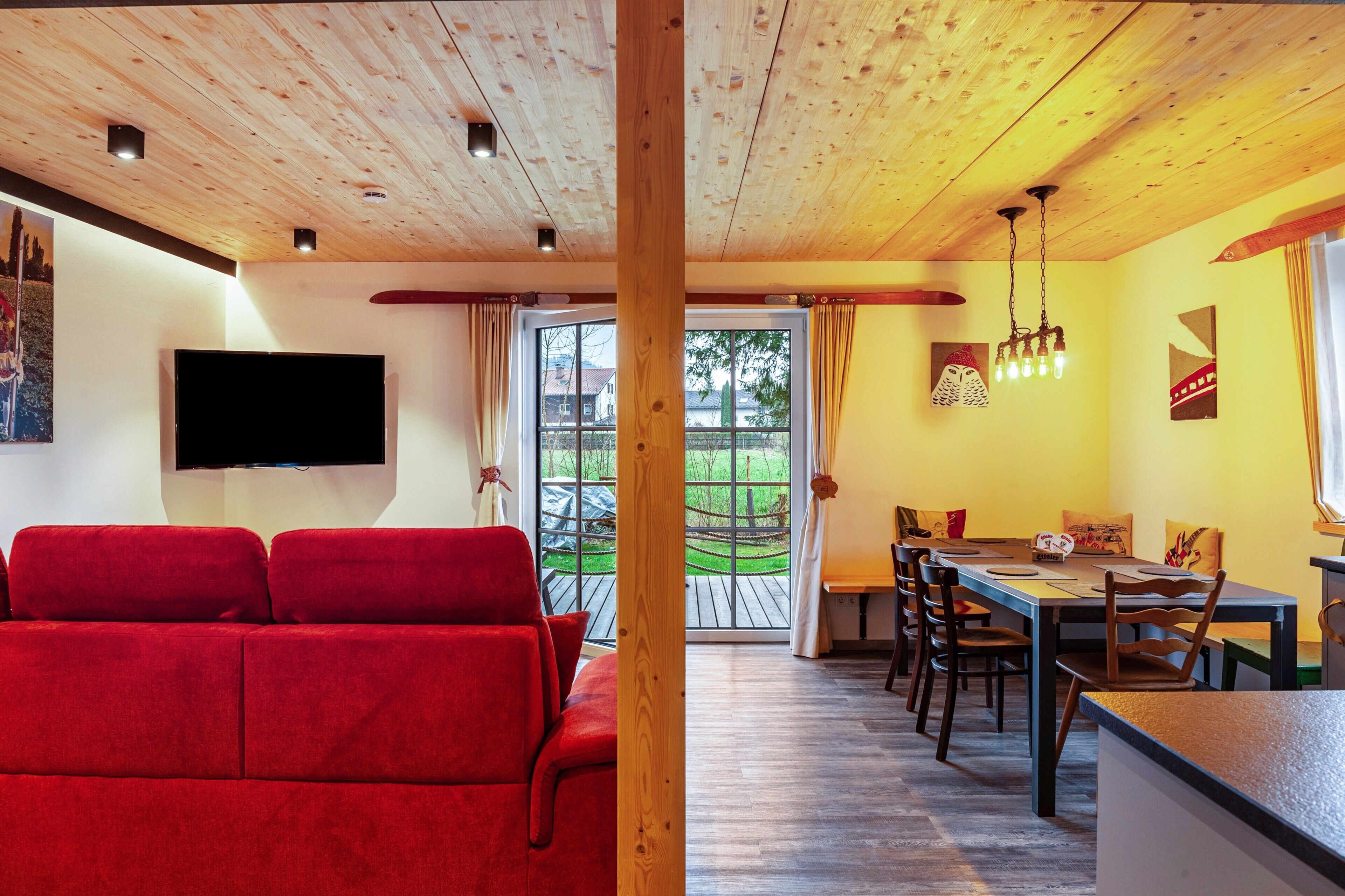 A room with a wooden ceiling, a red sofa, a dining table with chairs, and a TV mounted on the wall.