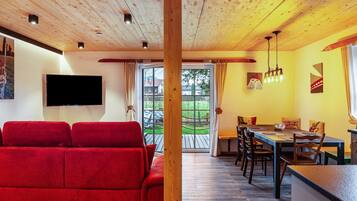 A room with a wooden ceiling, a red sofa, a dining table with chairs, and a TV mounted on the wall.