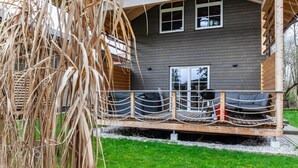 Une terrasse en bois avec une barrière en corde, une maison grise avec une fenêtre et une zone herbeuse.