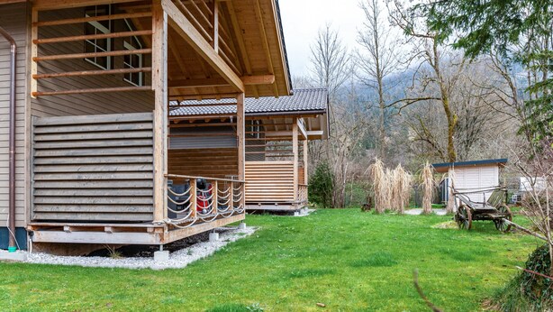 A wooden cabin with a porch, a cart, and a small shed in a grassy area with trees and mountains in the background. - Chalet Rauschn with mountain view with shared sauna, terrace, balcony; pet friendly (Farchant)