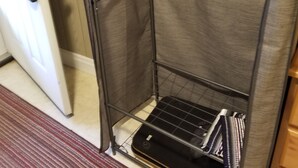 A corner of a room with a metal shelf unit containing folded clothes and a black object. A striped rug is on the floor.