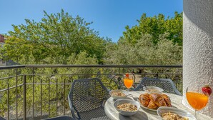 Ein Balkon mit Sitzgelegenheit im Freien, Frühstücksangeboten wie Croissants, Müsli und Orangensaft sowie Blick auf Bäume und Gebäude.