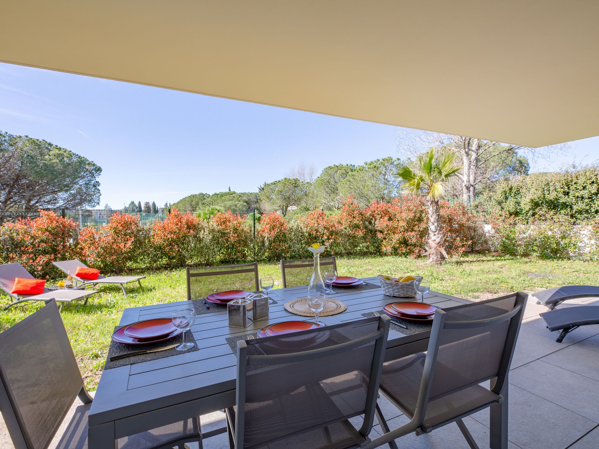 An outdoor dining area with a table set for two, surrounded by chairs, with a view of a garden and trees in the background.