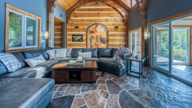 A log cabin interior with a stone floor, a large wooden ceiling, and a cozy seating area with a fireplace. - Large Grand Bend Getaway. Swim, Golf, Relax (South Huron (Munic))