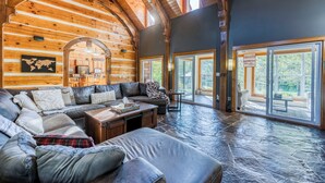 A spacious log cabin interior with a large living area, stone flooring, and large windows offering views of the outdoors. - Large Grand Bend Getaway. Swim, Golf, Relax (South Huron (Munic))