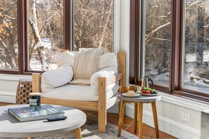 A cozy room with a white sofa, a small round table with a book, a remote, and a tray with fruits and a coffee pot.