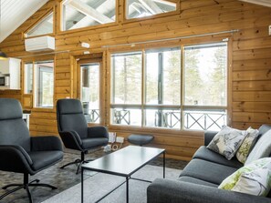A cozy cabin interior with a wooden interior, a sofa, two chairs, a small table, and large windows offering a view of the outdoors.