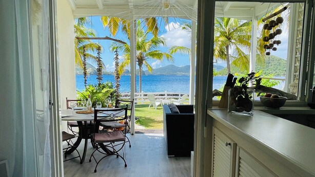 A room with a view of the sea, a dining table with chairs, and a kitchen area with a counter. - Blue Lagoon, Cottage feet in the water (Terre-de-Haut)