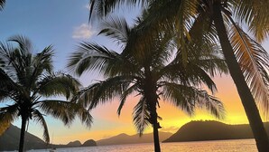 A beach with palm trees, a sunset, and boats on the water. - Blue Lagoon, Cottage feet in the water (Terre-de-Haut)