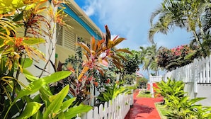 A pathway lined with white picket fences and lush tropical plants. - Blue Lagoon, Cottage feet in the water (Terre-de-Haut)