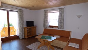 Een kamer met een houten vloer, een bank, een tafel met een groene placemat, een televisie op een houten kast, en een raam met gordijnen.