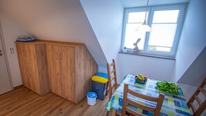 Una habitación pequeña y luminosa, con un armario de madera, una mesa de comedor con sillas, una ventana con persianas y una lámpara colgante.