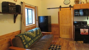 A cozy cabin interior with a wooden ceiling, a sofa, a television, a kitchen area with a stove and microwave, and a window with curtains.