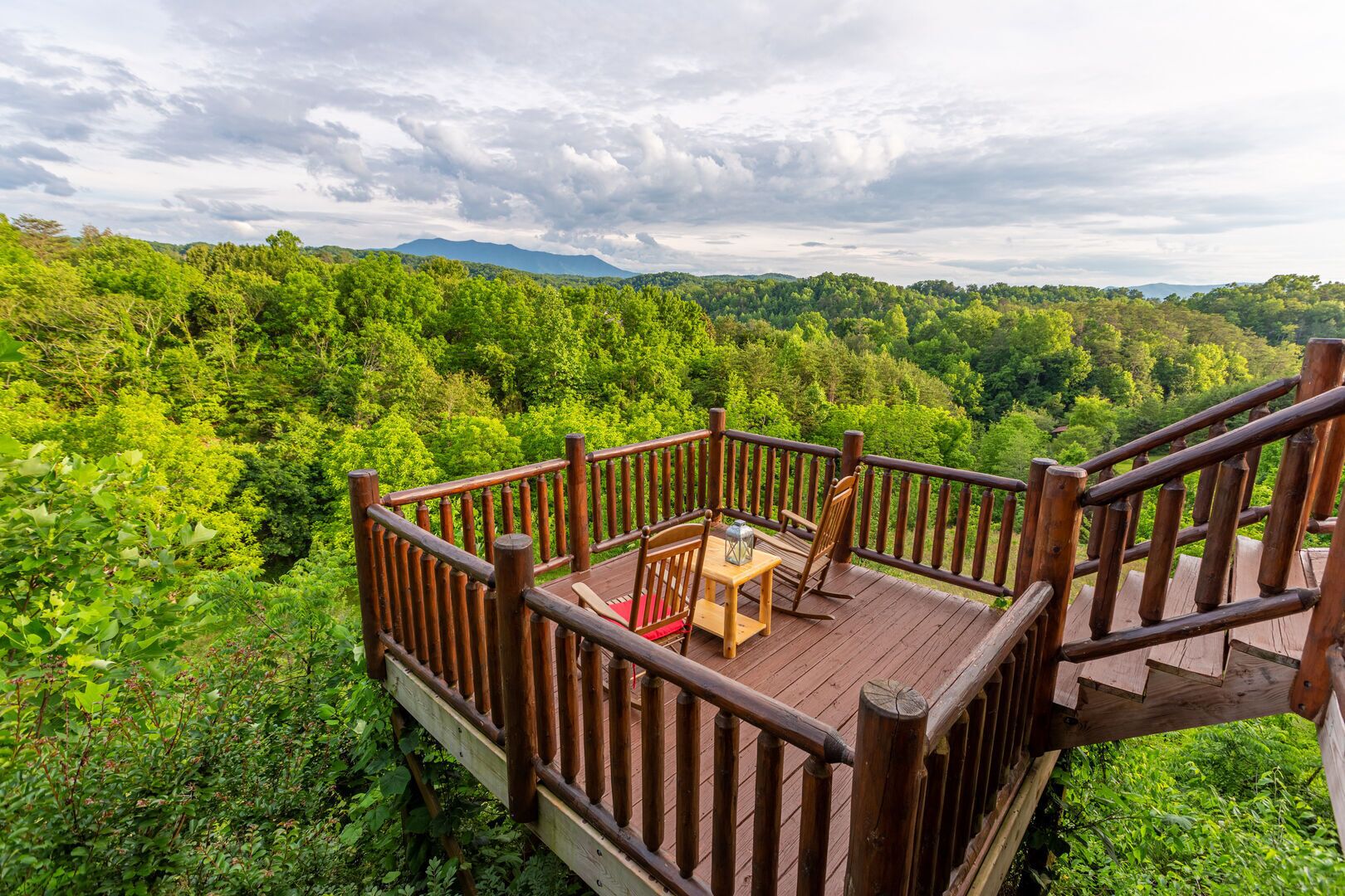 A wooden deck with a table and chairs, surrounded by lush greenery and a mountainous backdrop.