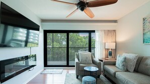 Ein modernes Wohnzimmer mit Flachbildfernseher, Deckenventilator, einer Couch, einem Sessel, einem kleinen runden Tisch und einem Balkon mit Blick auf Bäume.