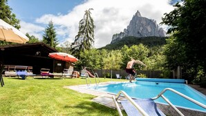 En swimmingpool med en bergskedja som bakgrund, omgiven av en gräsyta med solstolar och parasoller.