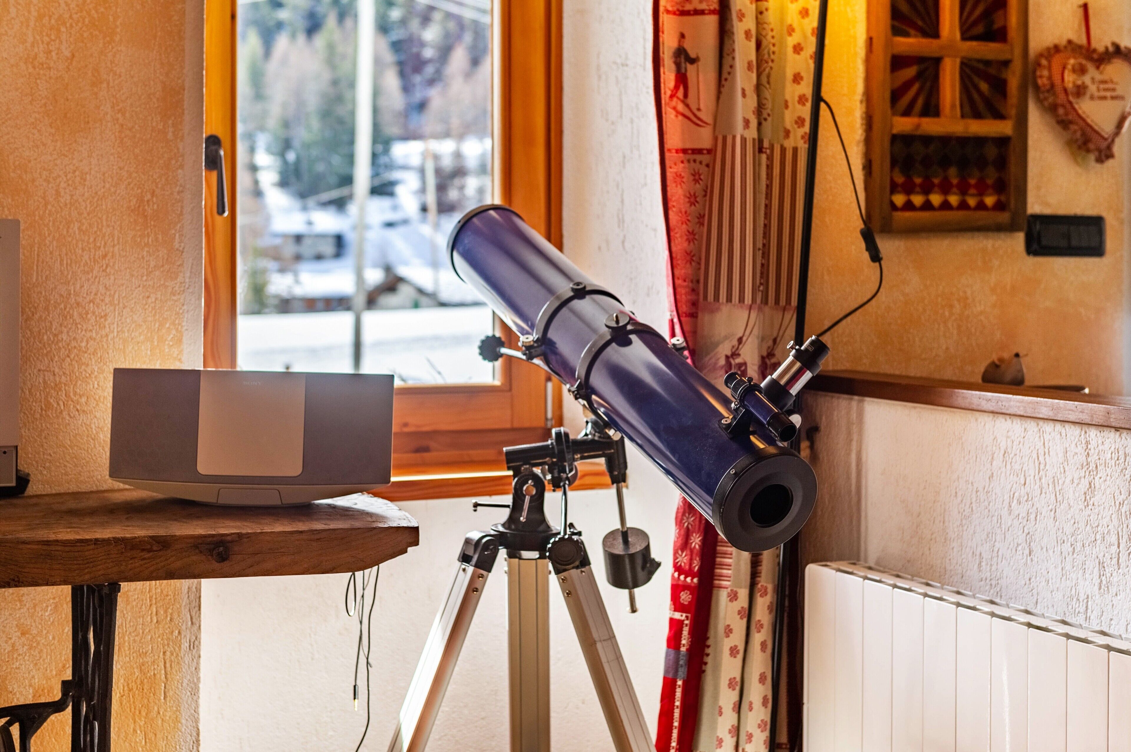 A telescope on a tripod in a room with a window, a radiator, and a wooden table.