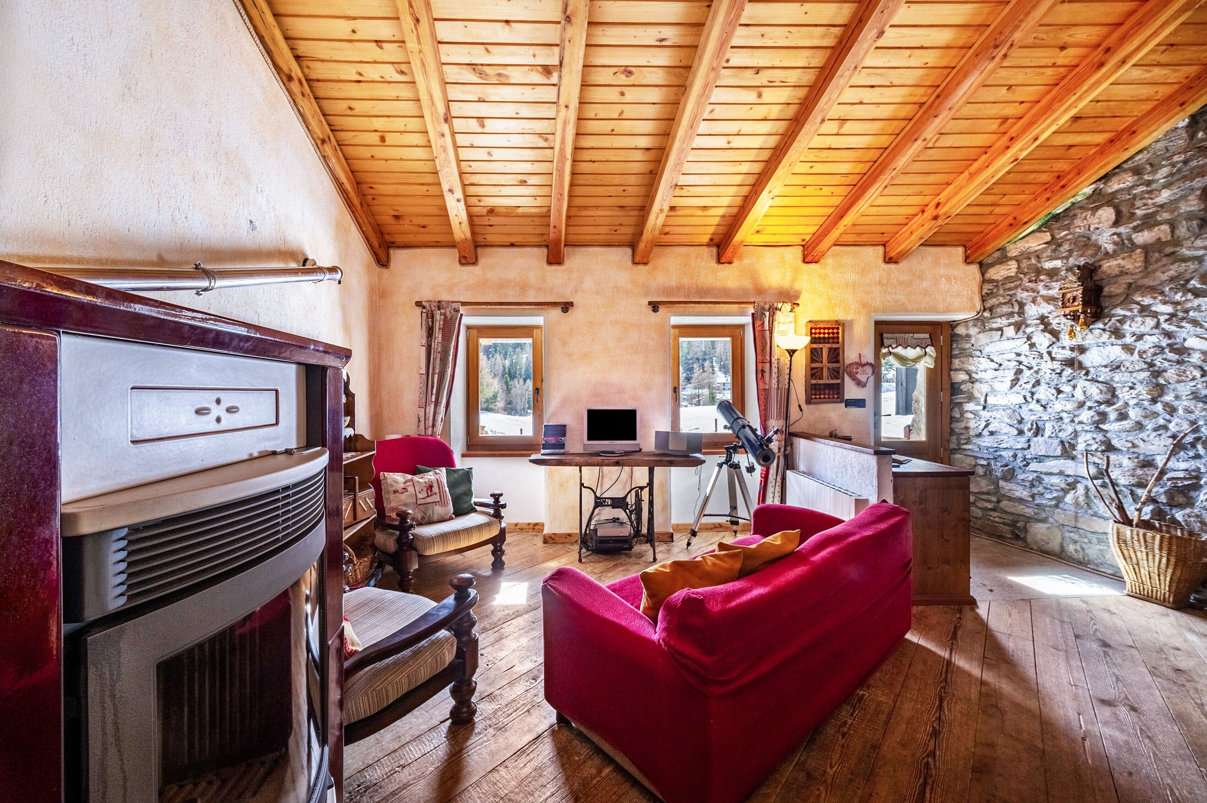 A cozy living room with a fireplace, wooden ceiling, stone wall, and a red sofa.