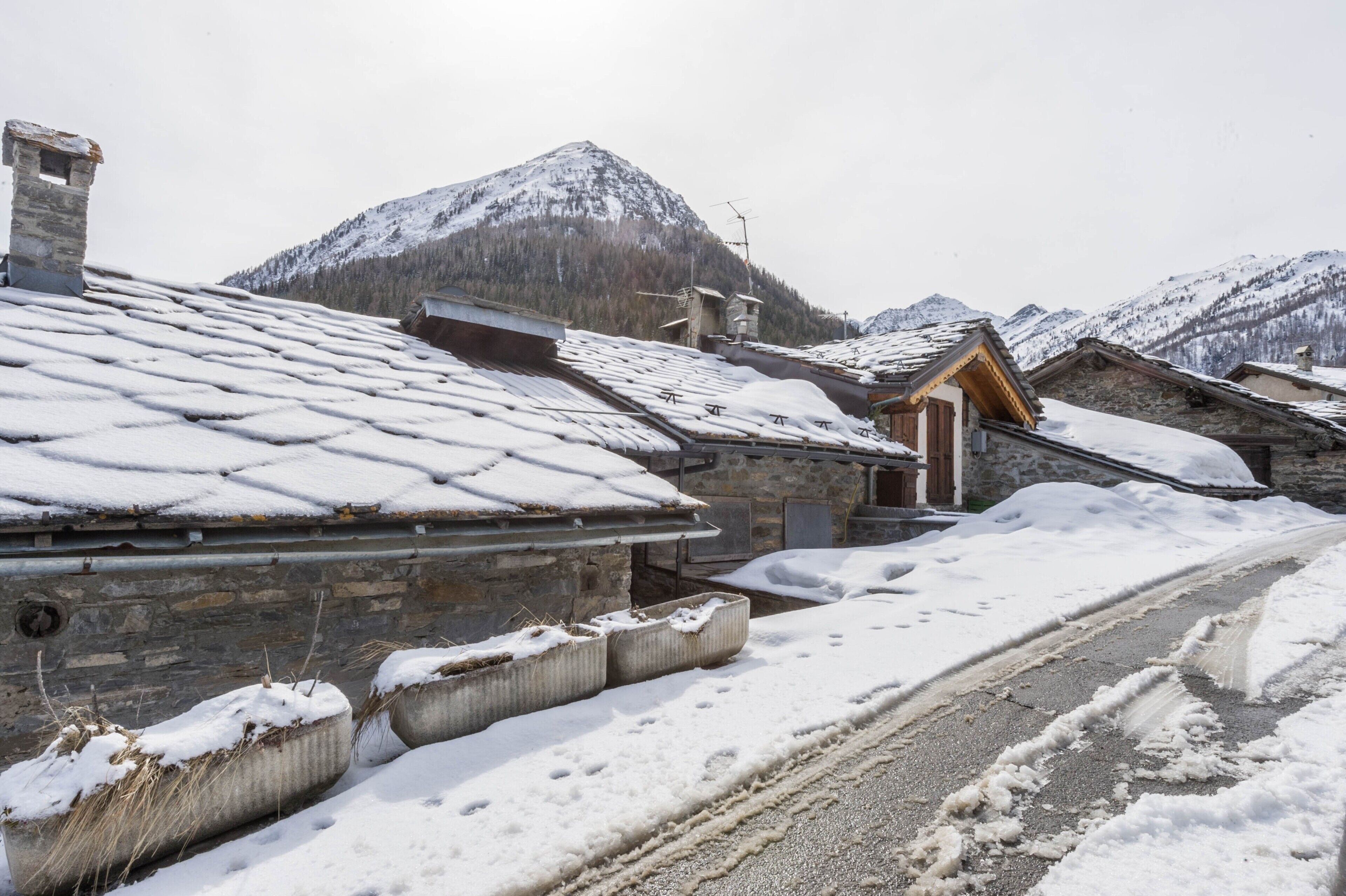 A snow-covered village with stone buildings, a road, and mountains in the background.