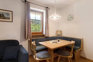 A room with a dining table, bench seating, a sofa, a window with curtains, a chandelier, and a wall clock.