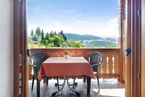 An open wooden door leading to a balcony with a table and chairs, overlooking a scenic landscape with trees and hills.