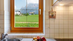 A kitchen with a window view of a green field and mountains, a stainless steel sink, and a fruit bowl on the counter. - Apartment "Stinerhof - Schlernblick" with Mountain View, Wi-Fi, Balcony & Garden (Völs am Schlern)