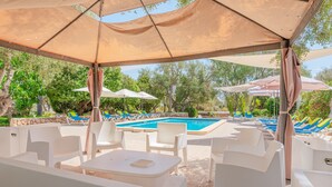 Ein überdachter Außenbereich mit weißen Stühlen und Tischen, einem Pool und Sonnenschirmen.
