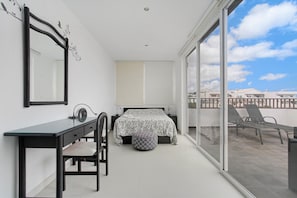 Un dormitorio con cama, escritorio, silla, ventana grande y balcón con sillas.