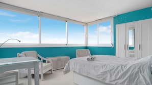 A bedroom with a bed, a desk, a chair, a laundry basket, and a window with a view of the sky.