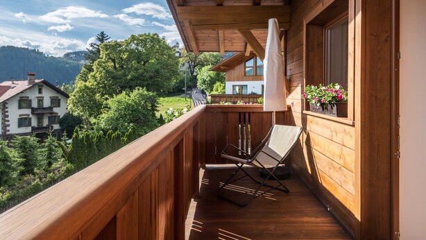A wooden balcony with a chair and umbrella, overlooking a mountainous area with houses and greenery. - Apartment 'Kristiania Type A2' with Mountain View, Sauna, Balcony & Garden (Siusi allo Sciliar (BZ))
