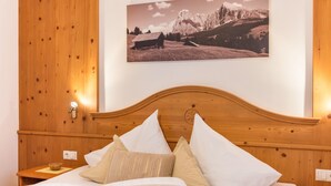 Ein Bett mit hölzernem Kopfteil, mit weißen und beigen Kissen, ein Nachttisch mit Lampe sowie ein Gemälde einer Berglandschaft an der Wand.