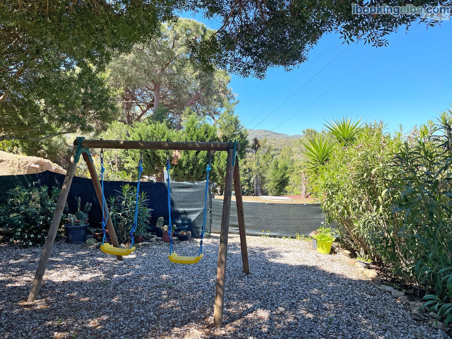 A wooden swing set with yellow seats in a garden area with gravel ground, surrounded by various plants and trees.