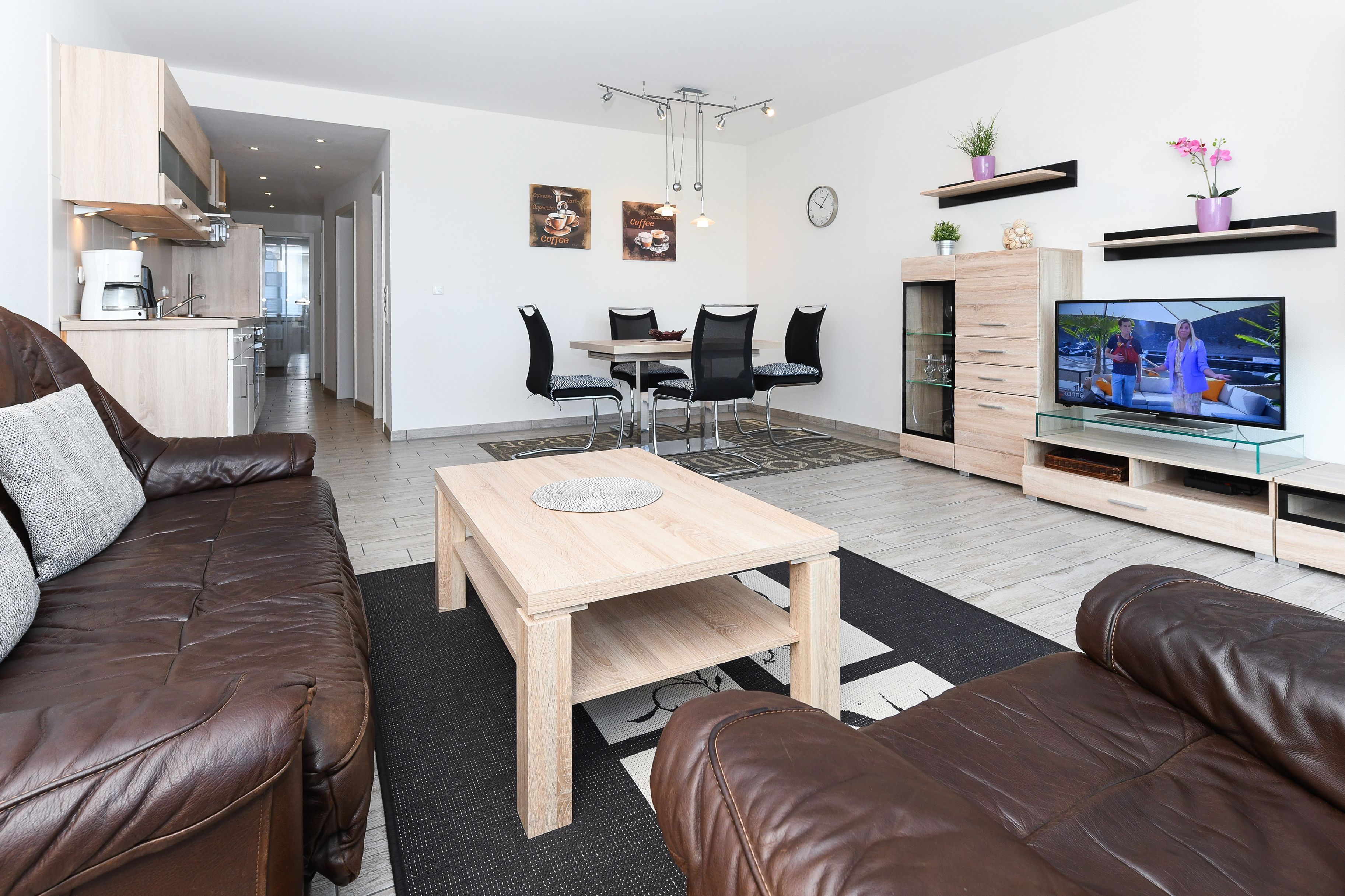 Un salon moderne avec un canapé en cuir marron, une table basse en bois et un téléviseur. On y trouve également un coin repas avec une table et des chaises, et une cuisine à l’arrière-plan.