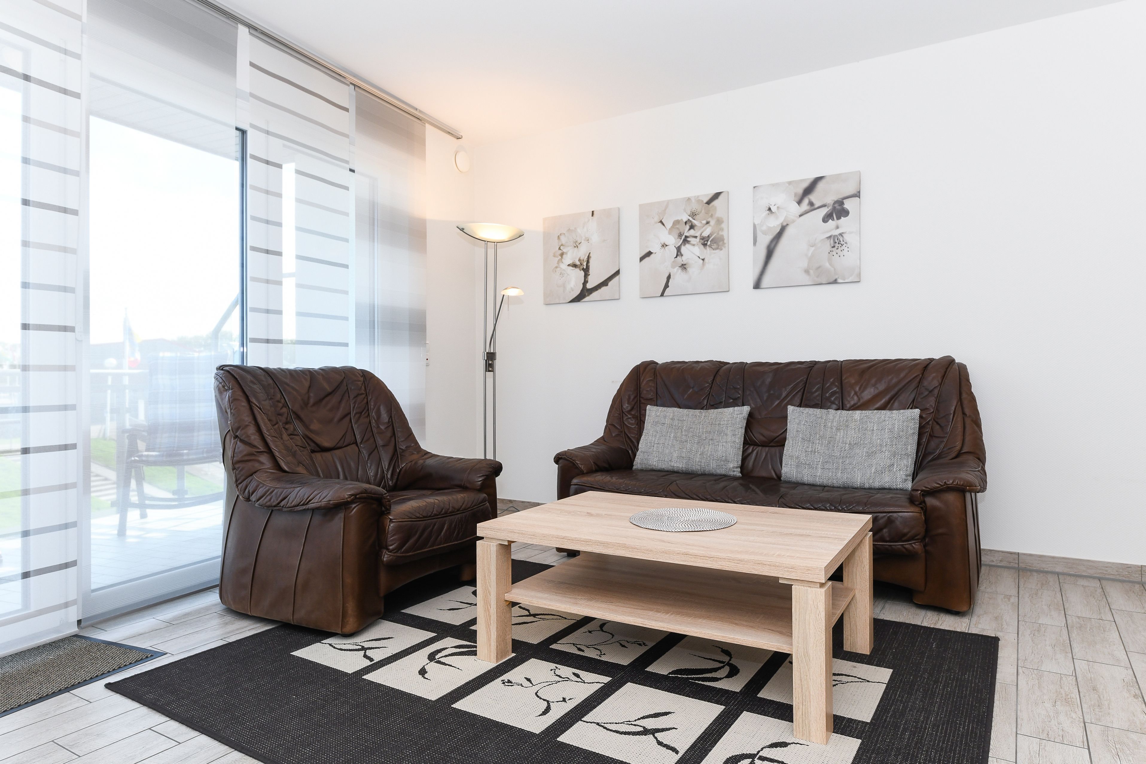 Un salon moderne avec un canapé en cuir marron, une table basse en bois et un tapis à motifs noir et blanc. Trois tableaux représentant des fleurs sont accrochés au mur, et une lampe sur pied est placée dans un coin.