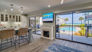 A modern kitchen with white cabinets, a dining area with wicker chairs, a fireplace, and a large TV.