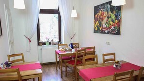 A dining area with wooden tables and chairs, a window with curtains, and a fruit still life painting on the wall.