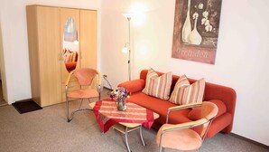 A room with a sofa, two chairs, a small table with a vase of flowers, a cabinet with a mirror, and a wall-mounted lamp.