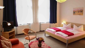 A hotel room with a bed, a TV, a chair, a table with a vase of flowers, and a painting on the wall.
